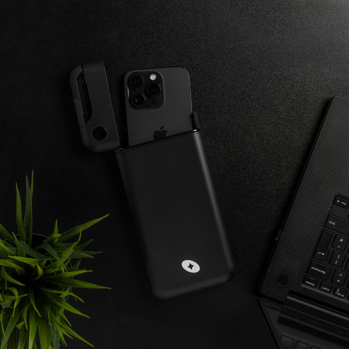 The Lockbox on a dark desk beside a plant and laptop, phone sliding into the box